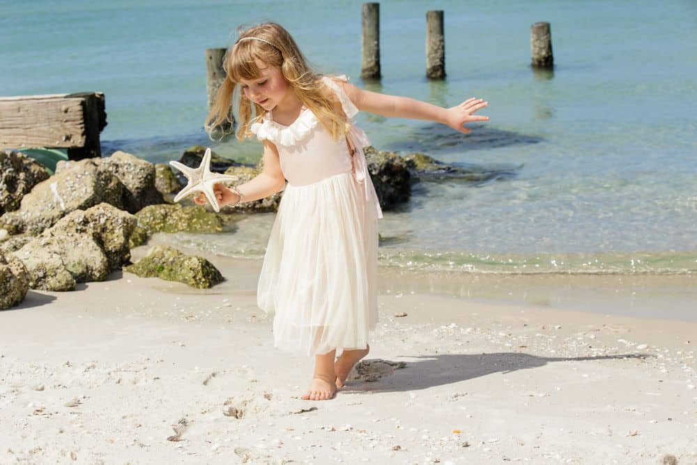 A young girl in a white dress holds a starfish while walking barefoot along a sandy beach, with rocks, calm blue water, and wooden posts in the background.