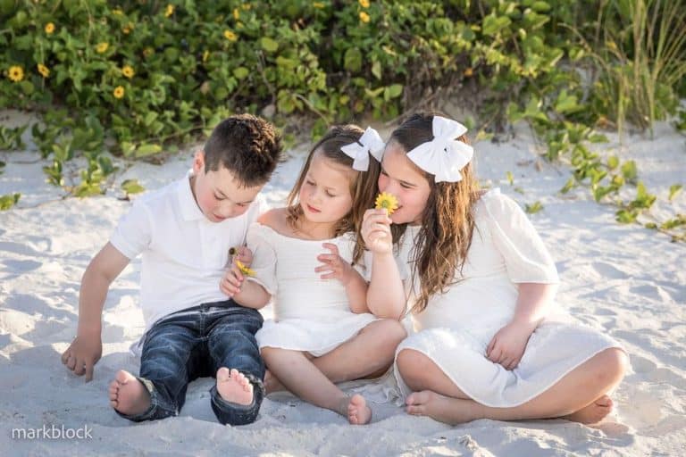 Three young children sit together on a sandy beach, all dressed in white. Two girls with white bows in their hair hold yellow flowers, while the boy looks down, sitting barefoot. Greenery is visible in the background.