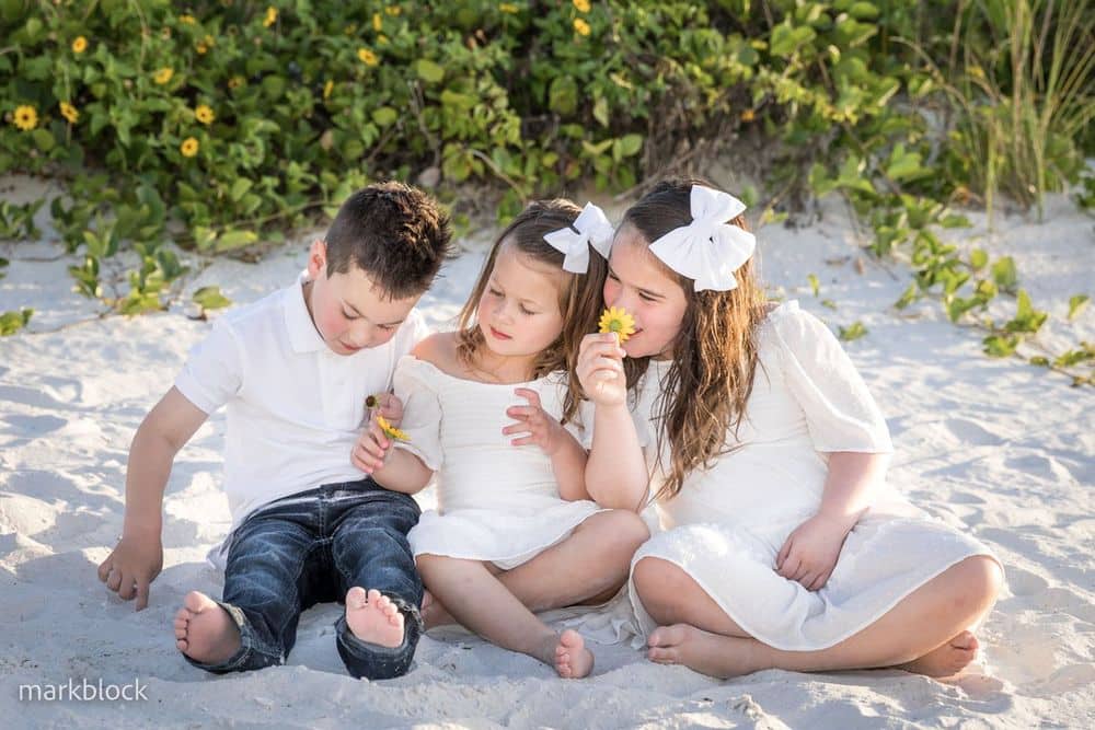 Three young children sit together on a sandy beach, all dressed in white. Two girls with white bows in their hair hold yellow flowers, while the boy looks down, sitting barefoot. Greenery is visible in the background.
