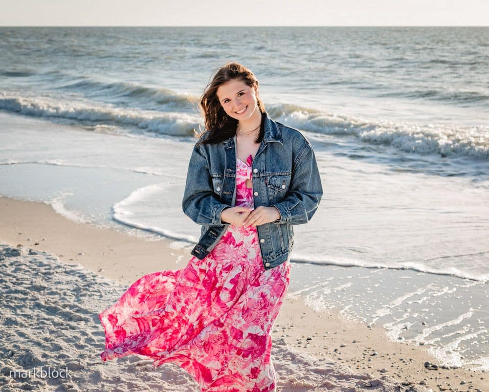 A young woman stands on a sandy beach by the ocean, wearing a pink and white floral dress with a denim jacket. She smiles at the camera, with gentle waves and sunlight in the background.