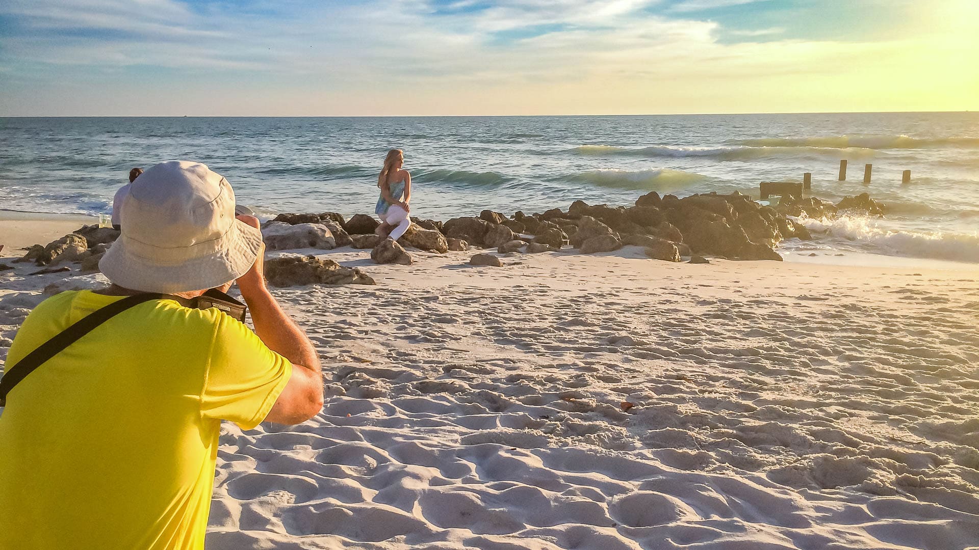 A Naples Marco Island photographer in a yellow shirt and white hat captures a woman posing on rocks by the ocean at a sandy beach during sunset, with partly cloudy skies and water reflecting the golden sunlight.