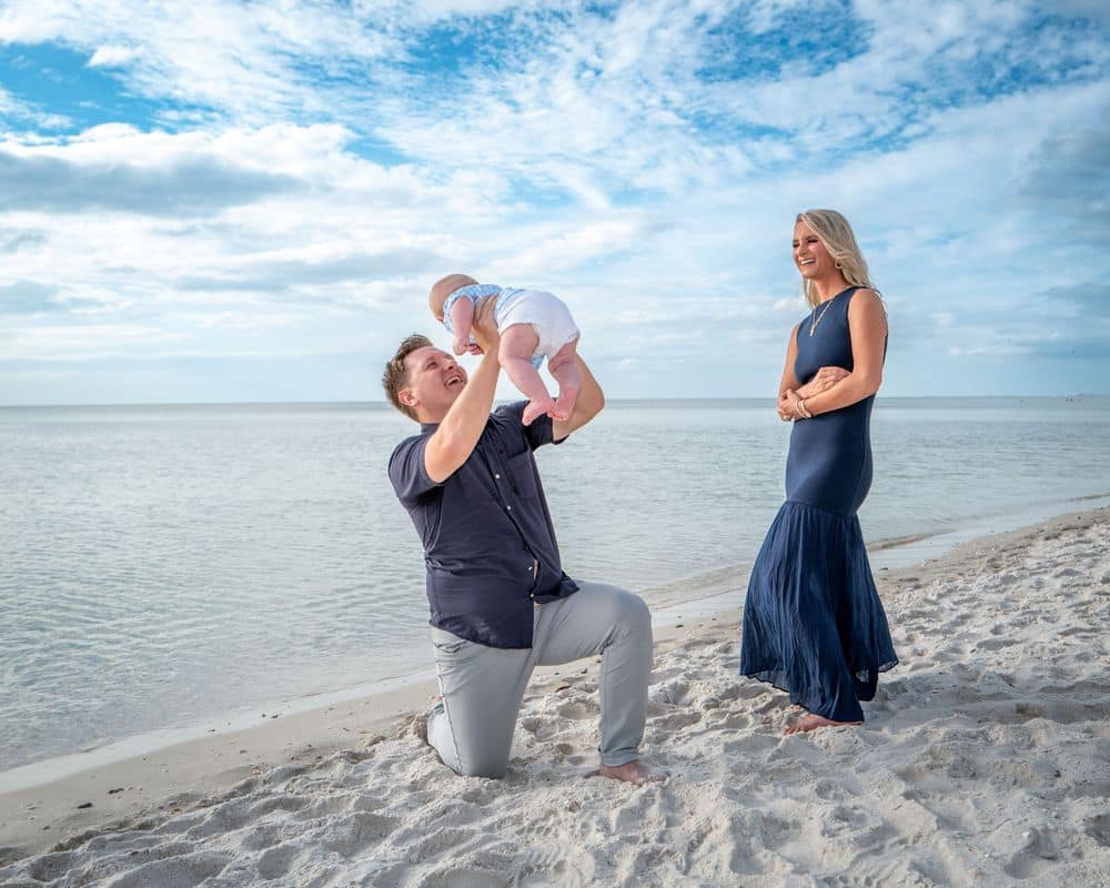 A man kneels on a sandy beach, lifting a baby in the air, while a woman in a navy dress stands nearby, smiling. The ocean and a partly cloudy sky are in the background.
