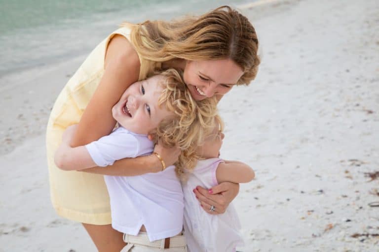 A woman in a yellow dress hugs and smiles with two young children on a sandy beach during family beach portraits. The children, both with blonde hair, are laughing and wearing light-colored clothes, with the ocean visible in the background.