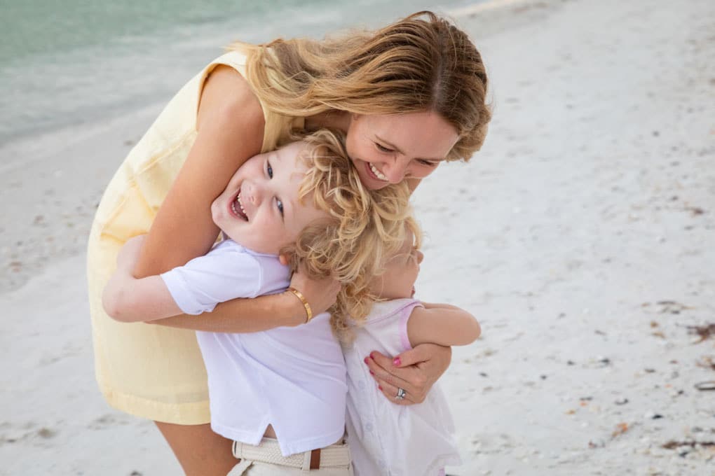 A woman in a yellow dress hugs and smiles with two young children on a sandy beach during family beach portraits. The children, both with blonde hair, are laughing and wearing light-colored clothes, with the ocean visible in the background.