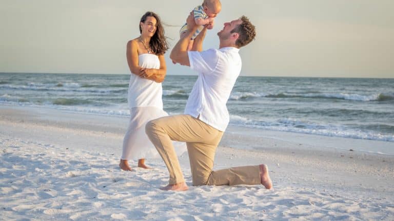 A man kneels on the beach, holding a baby up and smiling, while a woman in a white dress stands nearby, watching happily—capturing a perfect moment that shows how to plan beach family portraits with natural joy and scenic ocean waves.