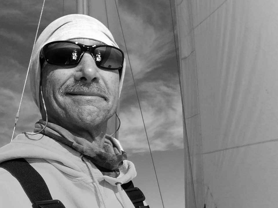 A person wearing a hat, sunglasses, and a jacket smiles while sailing. Captured by a Naples Marco Island photographer, the black and white image features large sails and a partly cloudy sky in the background.