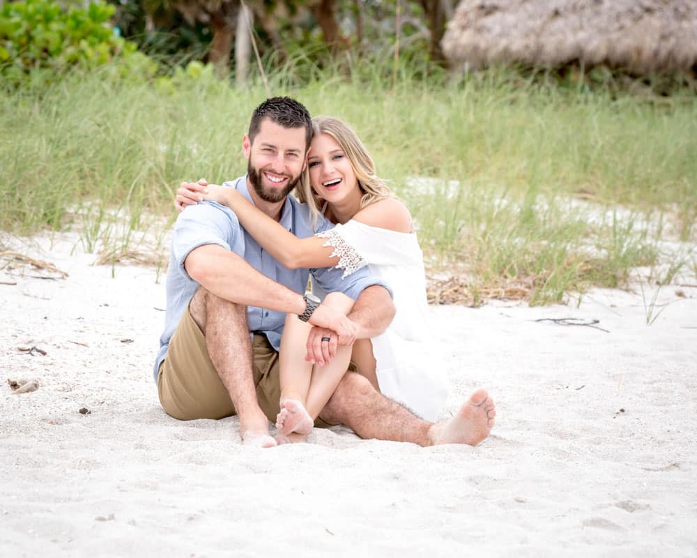 A smiling couple sits close together on a sandy beach, surrounded by greenery. The woman has her arm around the mans shoulder, and they both look happy and relaxed.