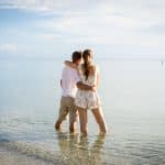 A couple stands in shallow, clear water at the edge of the sea, embracing and looking out toward the calm horizon under a partly cloudy sky.