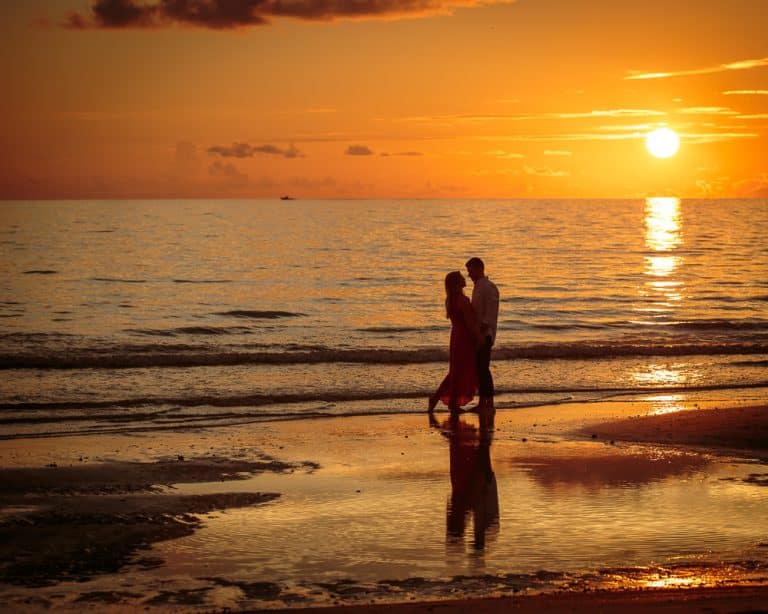 A couple walks along the beach at sunset, holding hands with the sun low in the sky, casting an orange glow over the water and sand.