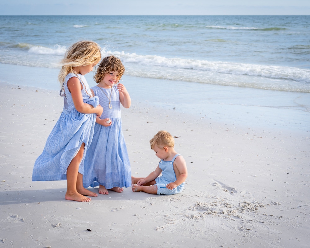 Two young girls in blue dresses stand on a sandy beach near the ocean, smiling and looking at a toddler boy in blue overalls who is sitting and playing in the sand. Waves and sky are visible in the background.