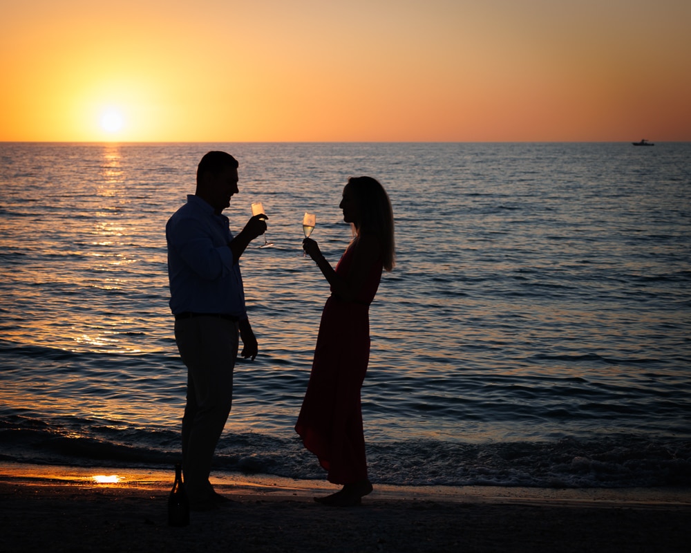 Silhouette of a man and woman toasting with drinks on a beach at sunset, with calm waves and the sun low on the horizon.