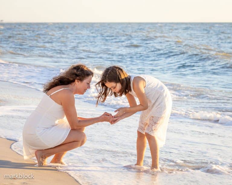 A woman and a young girl, both in white dresses, crouch together at the edge of the beach, smiling as they examine something in the girls hands. Waves gently lap at their feet under a clear sky.