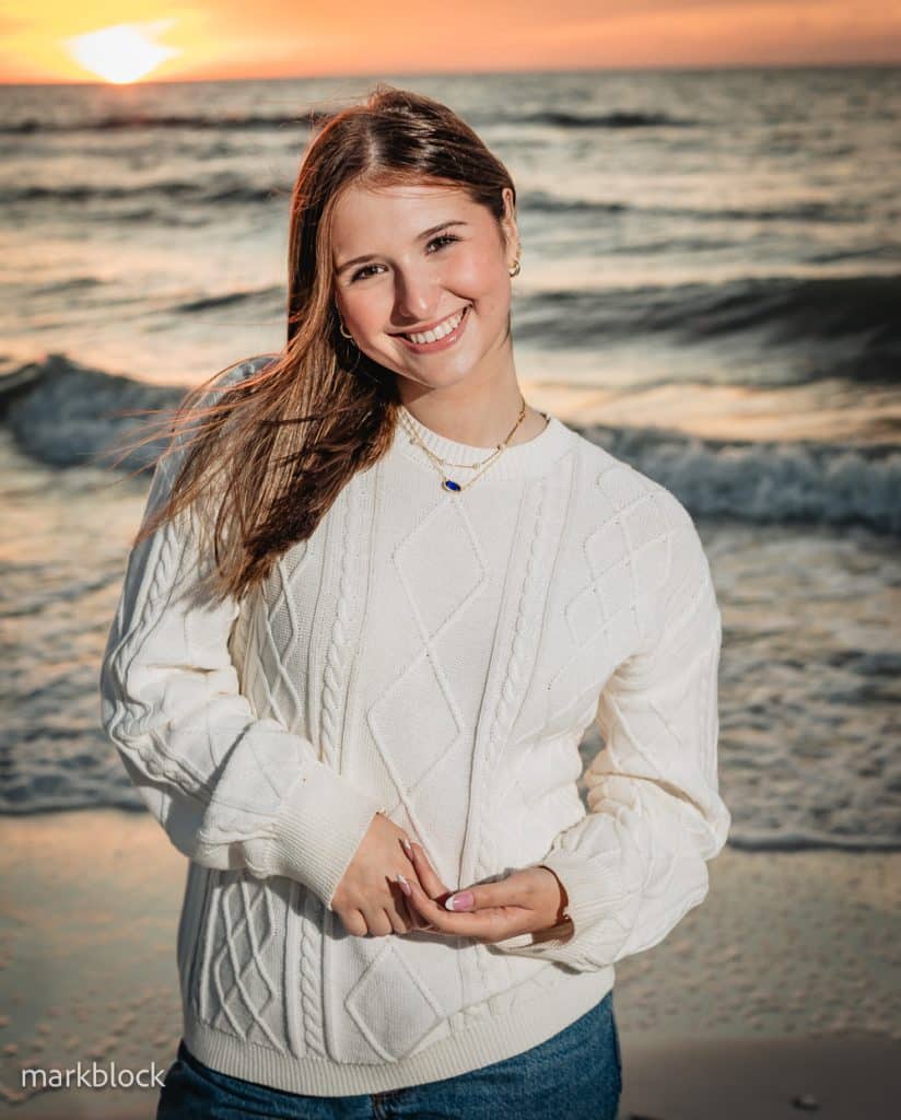 A young woman in a white sweater smiles while standing on a beach at sunset, with waves and the orange sun visible in the background.