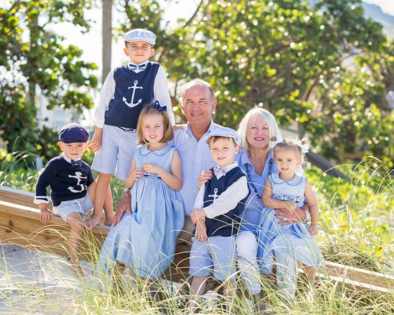A smiling older couple sits on a wooden fence outdoors with four young children and one older child, all dressed in blue and white outfits. Captured by a Naples family beach photographer, the lush green foliage and sunlight create a warm, joyful scene.