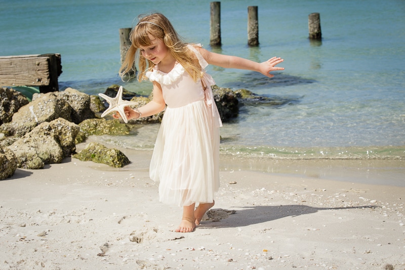 A young girl in a white dress walks barefoot on a sandy beach, holding a starfish. Captured by a Naples beach portrait photographer, rocks and weathered wooden posts sit near the clear, blue ocean in the background.
