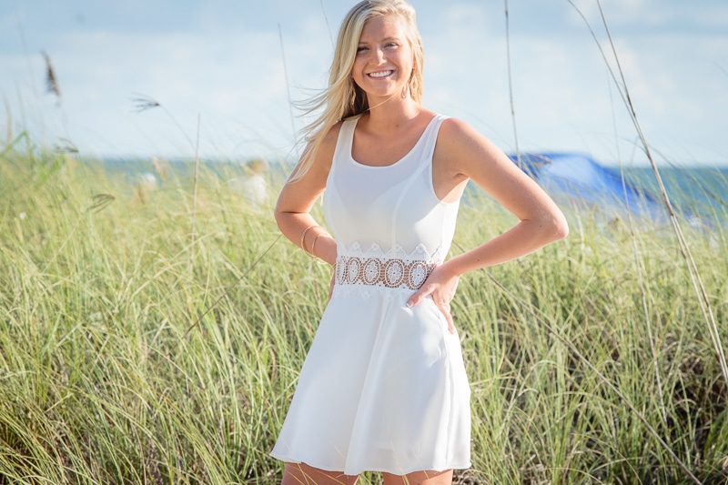 A woman in a sleeveless white dress stands smiling with her hands on her hips in a grassy area near the beach, with the ocean and blue sky behind her—capturing the perfect moment for a Naples beach portrait photographer.