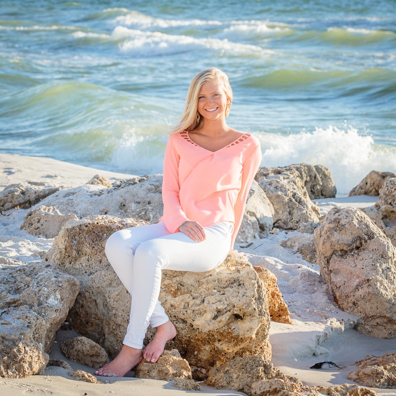 A smiling woman in a light pink top and white pants sits barefoot on large rocks at the beach, with waves and the ocean in the background under a bright sky, captured by a Naples beach portrait photographer.