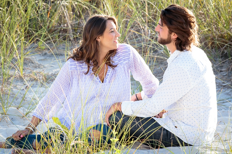 A woman and a man sit together on sandy ground among tall grass, facing each other and smiling warmly. Sunlight highlights their relaxed outfits as a Naples beach portrait photographer captures this serene outdoor moment.