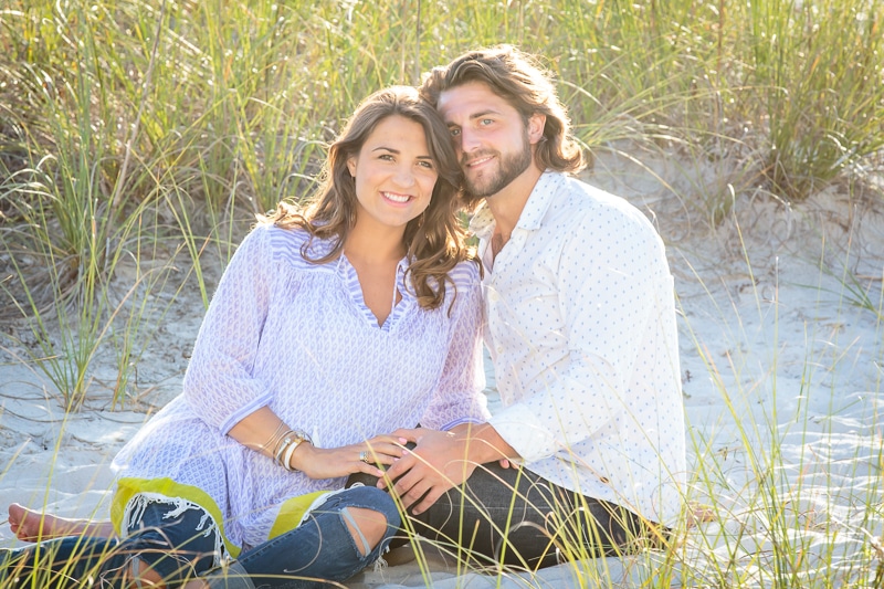 A smiling couple sits close together on sandy ground surrounded by tall beach grass, both wearing light-colored clothes and looking happy in the sunlight—a picture-perfect moment captured by a Naples beach portrait photographer.