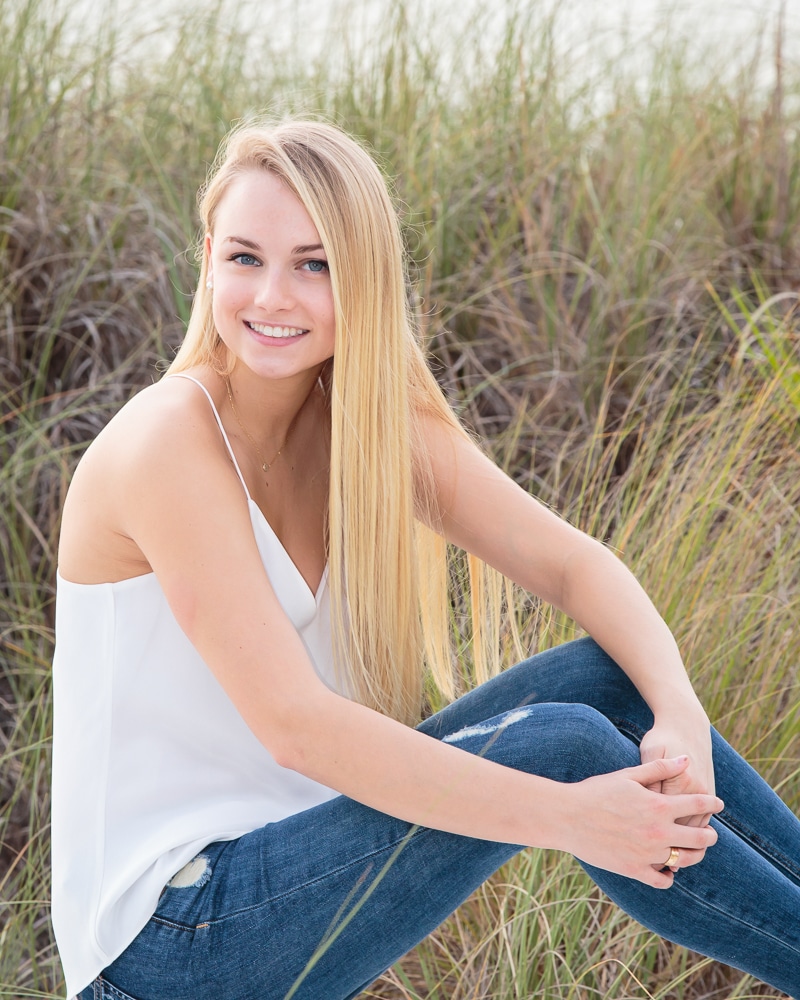 A young woman with long blonde hair, wearing a white sleeveless top and blue jeans, sits on the grass outdoors, smiling at the camera—captured in natural light by a Naples beach portrait photographer. Tall green grass is visible in the background.