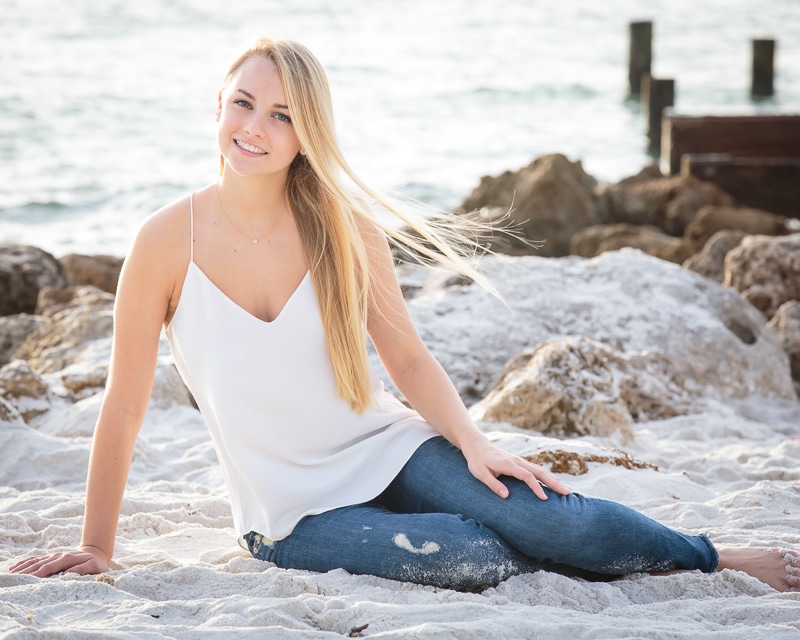 A young woman with long blonde hair, wearing a white sleeveless top and blue jeans, sits on the sandy beach near rocks, smiling at the camera with the ocean in the background—captured by a Naples beach portrait photographer.