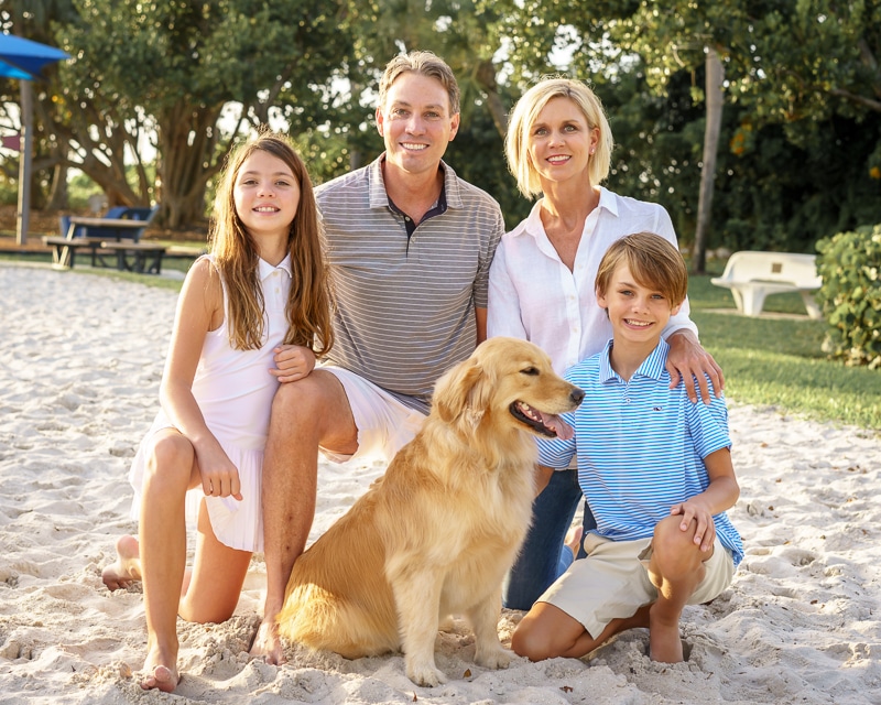 A smiling family of four and their golden retriever pose together on a sandy beach, with trees and picnic tables in the background—captured perfectly by a Naples beach portrait photographer. Parents stand behind their kids, who kneel beside the dog.