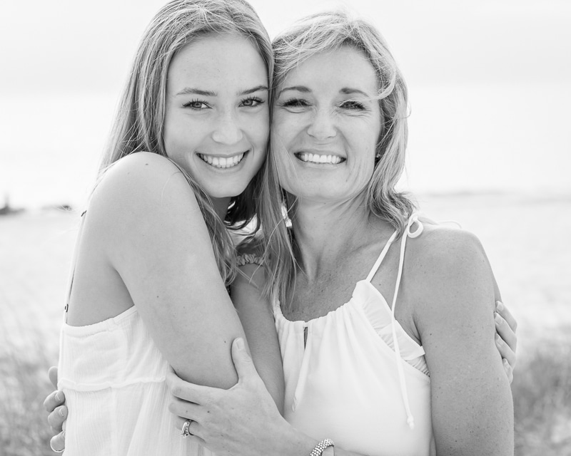 Two women, likely a mother and daughter, smile and embrace outdoors in this black and white image by a Naples beach portrait photographer. Both wear light sleeveless tops with the blurred beach scene softly visible behind them.