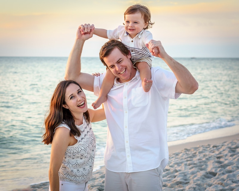 A smiling family of three stands on a sandy beach at sunset, captured by a Naples beach portrait photographer. The father holds a child on his shoulders, while the mother stands beside them in light summer clothes, with the ocean behind them.
