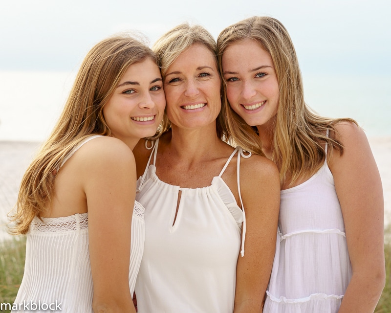 Three women, likely a mother and two daughters, stand close together on a beach, smiling at the camera. Captured by a Naples beach portrait photographer, they wear light summer clothes with the ocean visible in the background.