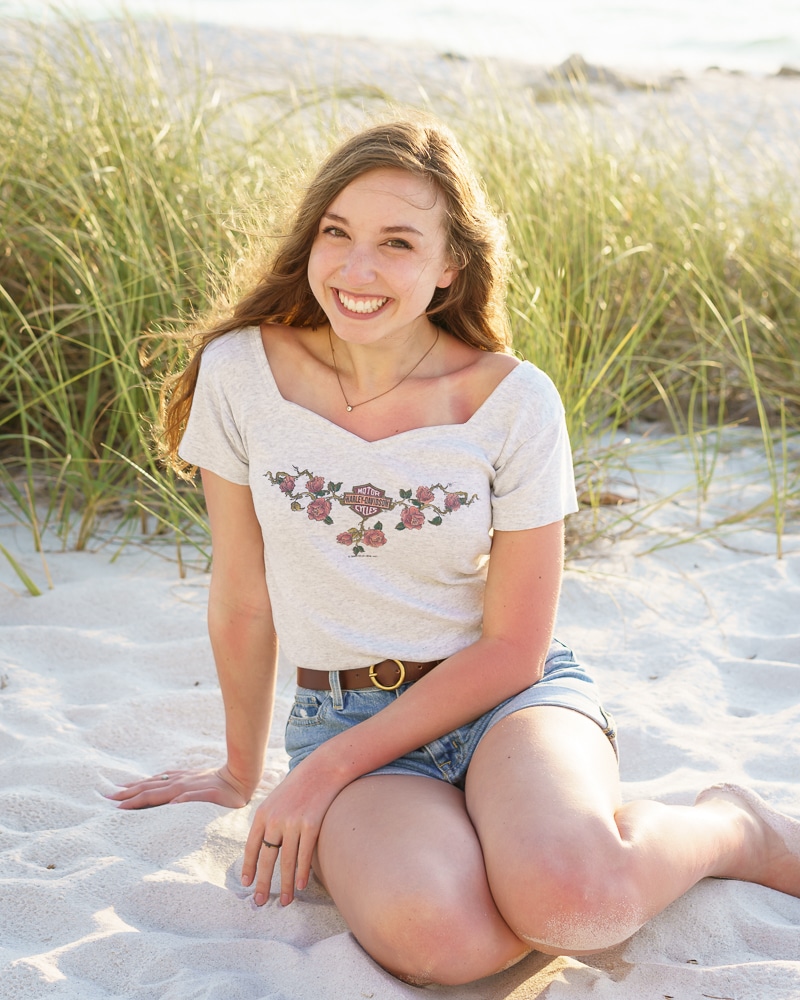 A young woman with long brown hair sits on sandy beach, smiling at the camera. Captured by a Naples beach portrait photographer, she wears a light gray floral shirt and denim shorts. Dune grass and bright sunlight are in the background.