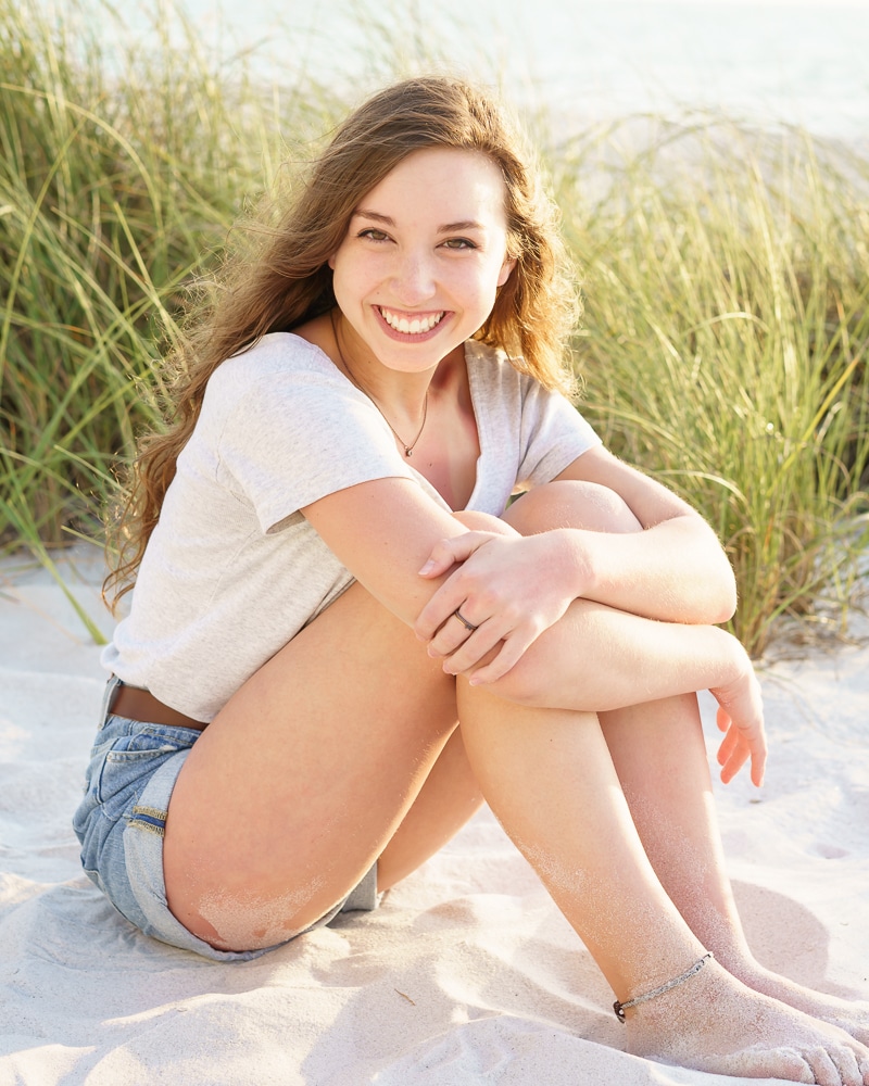 A young woman with long, wavy hair sits on sandy beach with grass in the background, smiling at the camera. Captured by a Naples beach portrait photographer, she wears a light gray t-shirt and denim shorts, her arms wrapped around her knees.