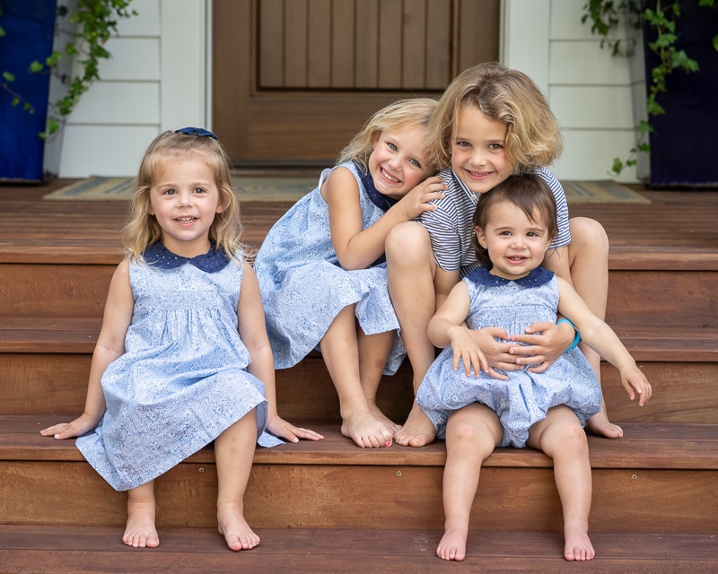 Four young children sit barefoot on wooden steps, smiling in matching blue outfits. Three girls huddle together while the youngest, also in blue, sits in an older child’s lap—captured beautifully by a Naples beach portrait photographer. A wooden door is behind them.
