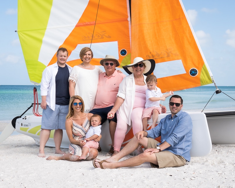 A multigenerational family poses together on a sandy beach in front of a sailboat with bright orange and yellow sails. Captured by a Naples beach portrait photographer, the group smiles in light summer clothing against the ocean and blue sky backdrop.