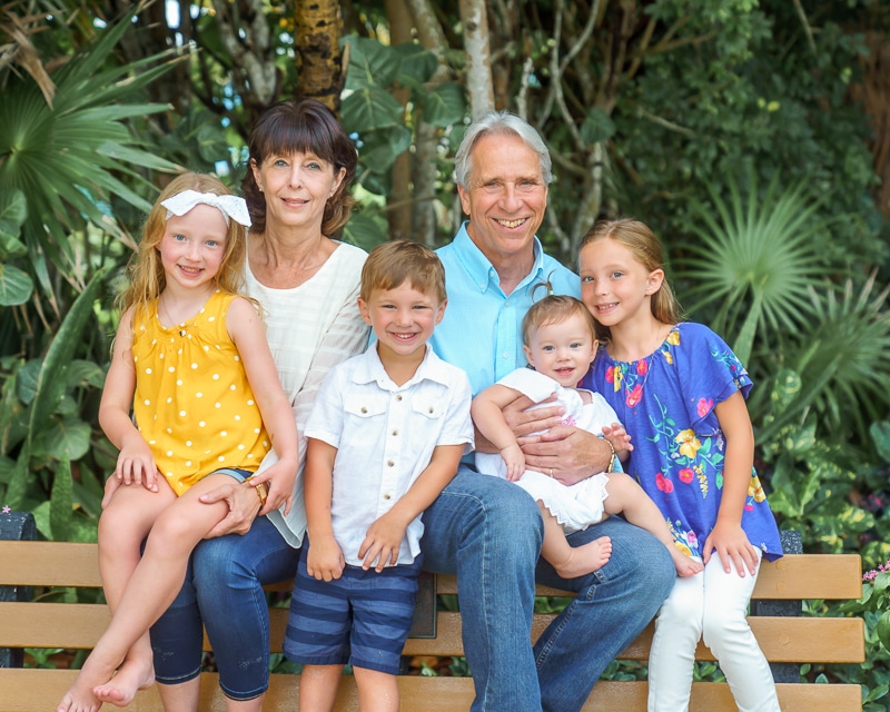 An older couple sits on a bench outdoors, surrounded by lush greenery, as four smiling young children—three girls and one boy—pose happily with them, captured beautifully by a Naples beach portrait photographer.