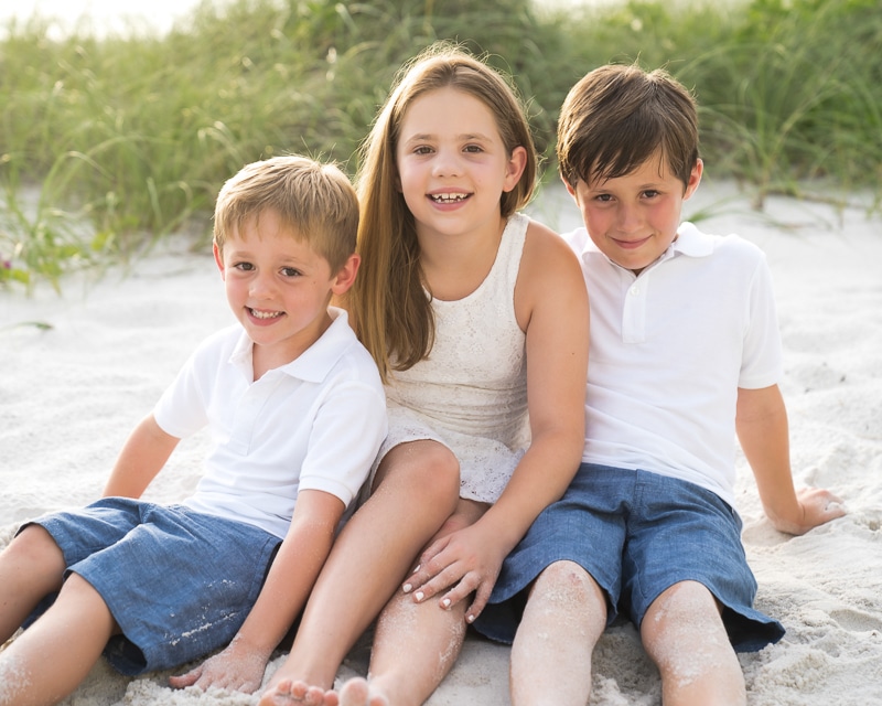 Three smiling children sit on the sand at the beach. Captured by a Naples beach portrait photographer, two boys in white shirts and blue shorts flank a girl in a white dress, with green plants visible in the background.
