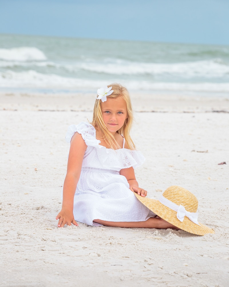 A young girl in a white dress with a flower in her hair sits on a sandy beach near the ocean, captured beautifully by a Naples beach portrait photographer. A straw hat with a white ribbon rests beside her as waves roll under the cloudy sky.