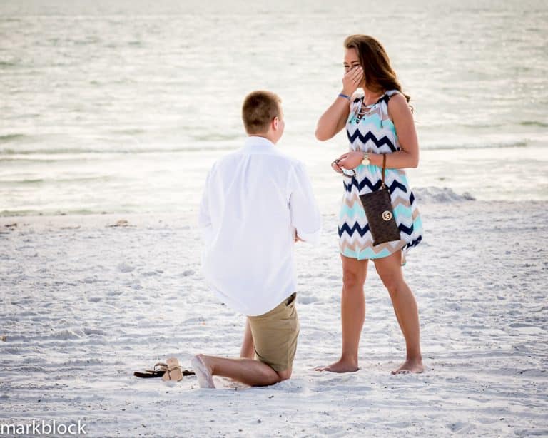 A man kneels on a sandy beach, proposing to a surprised woman who covers her mouth with her hand. The ocean glistens in the background—captured beautifully by a Naples beach portrait photographer as both appear emotional and happy.