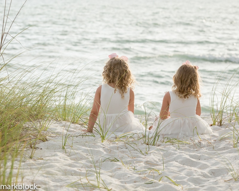 Two young girls in white dresses sit on sandy dunes, facing the ocean. Tall grasses surround them as they look out at the calm water—an idyllic moment captured by a Naples beach portrait photographer.