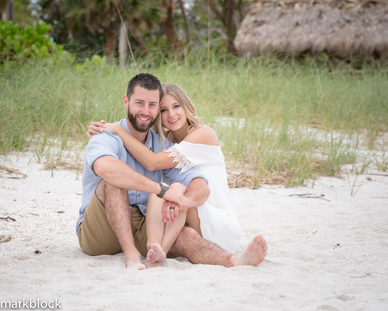 A smiling couple sits barefoot on sandy ground, with greenery and a thatched-roof structure in the background. Captured by a Naples beach portrait photographer, the woman leans into the man, both appearing relaxed and happy.