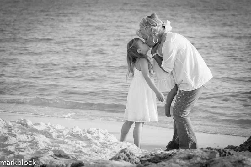 A woman and a young girl, both in light clothing, share a kiss on a sandy beach near the water’s edge. Captured in black and white by a Naples beach portrait photographer.