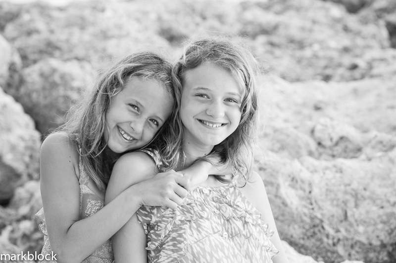 Two young girls smiling and hugging each other while sitting on rocky terrain. Captured in black and white by a Naples beach portrait photographer.