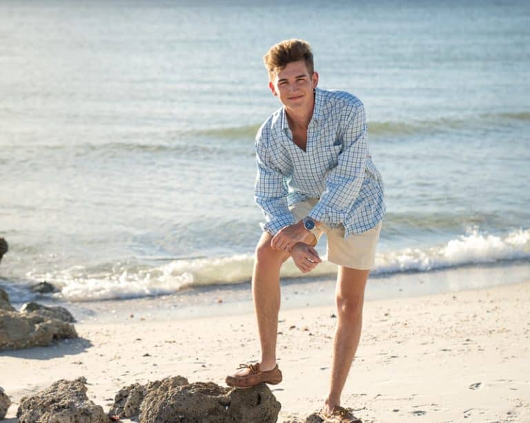 A young man in a light blue plaid shirt, khaki shorts, and boat shoes poses on a sandy beach near the water—an ideal scene for a Naples beach portrait photographer—with one knee bent, rocky shoreline, and calm waves in the background.