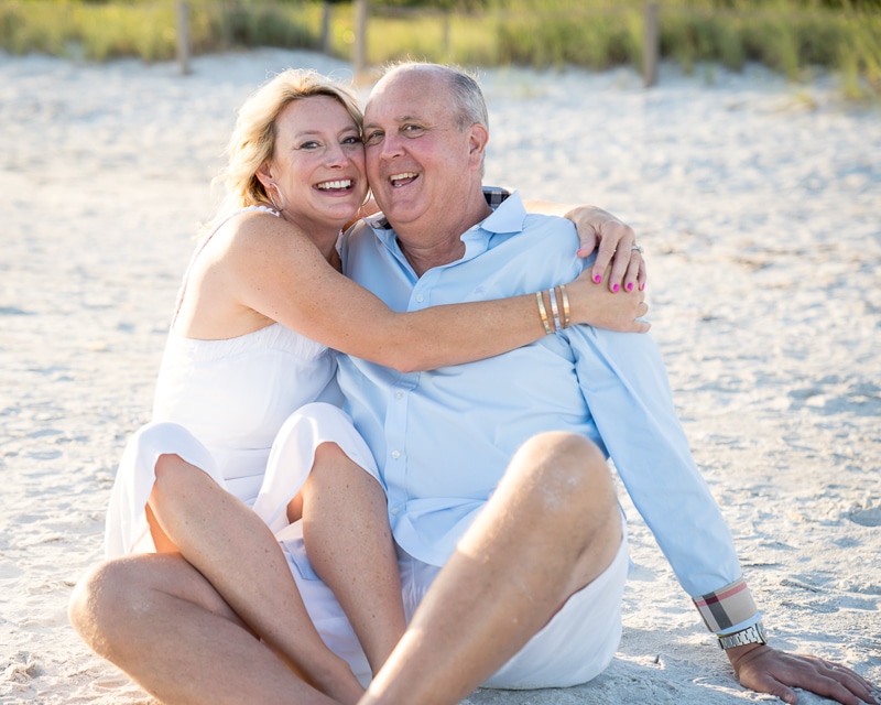 A smiling couple sits on sandy beach in casual clothes, the woman in a white dress hugging the man in a light blue shirt. Captured by a Naples beach portrait photographer, they appear happy and relaxed amid sunlit sand and beach grass.
