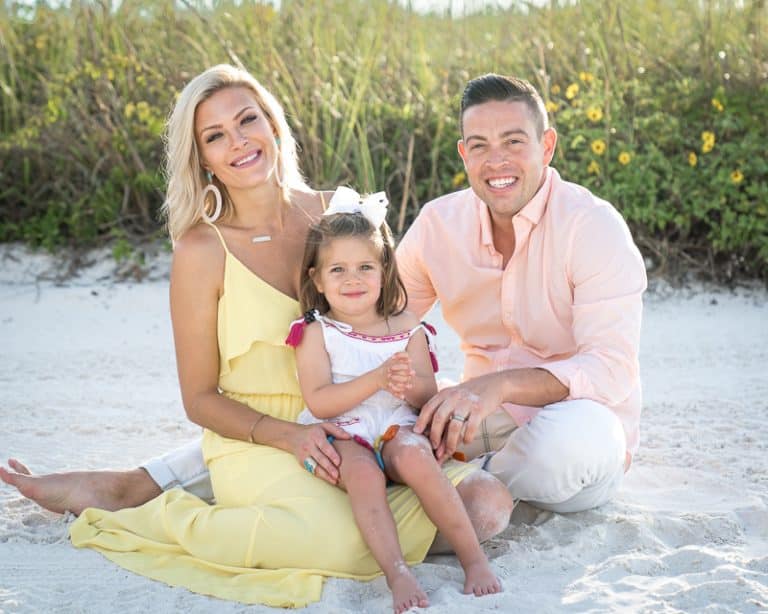 A smiling family of three sits on the sandy beach in front of tall grass, captured by a Naples beach portrait photographer. The mother wears yellow, the father light pink, and their young daughter a white dress with a bow in her hair.