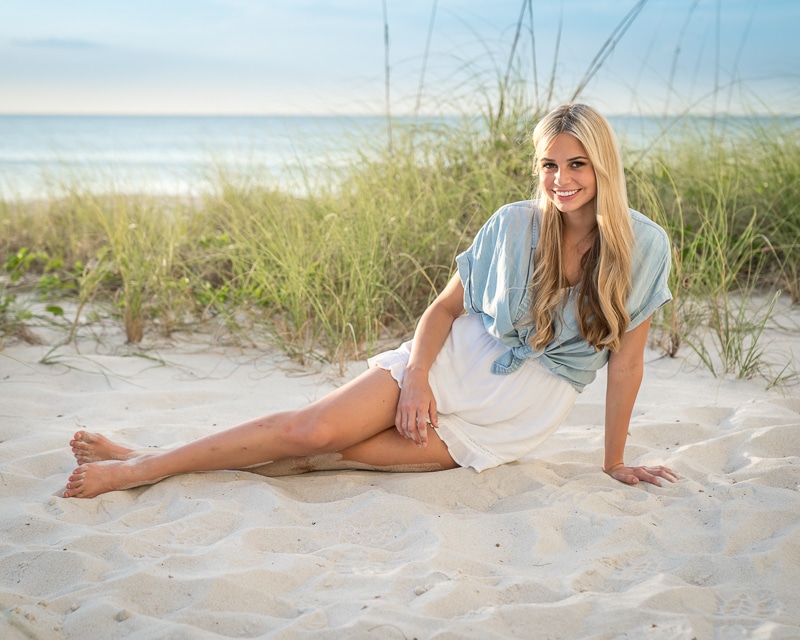 A young woman with long blonde hair sits on sandy beach, smiling at the camera. Captured by a Naples beach portrait photographer, she wears a light blue top and white skirt, with sea grass and the ocean in the background under a clear sky.
