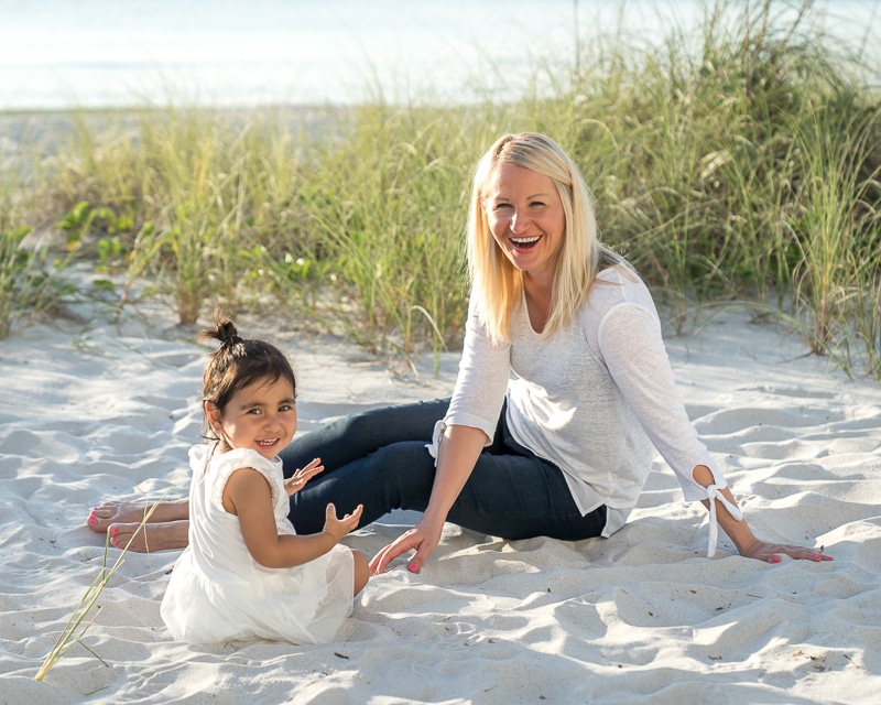 A woman and a young girl sit and smile on a sandy beach with grass in the background, beautifully captured by a Naples beach portrait photographer. The woman has blonde hair and wears a white top and jeans; the girl wears a white dress with a ponytail.