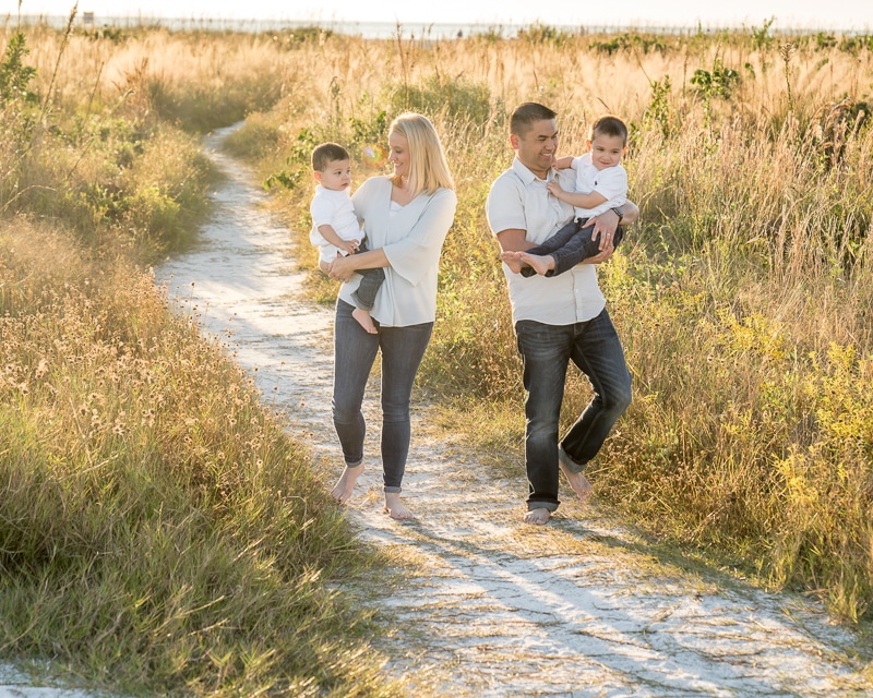A family of four walks barefoot on a sandy path through tall grass, sunlit and peaceful. Captured in the warm glow by a Naples beach portrait photographer, the parents in white tops and jeans each carry a young child, enjoying a perfect day.