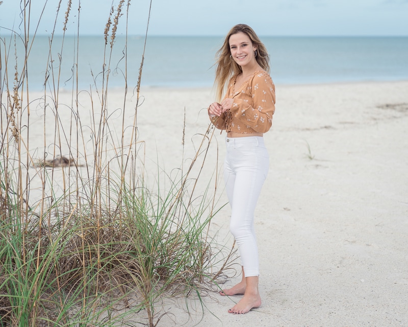 A young woman with long hair stands barefoot on a sandy beach near dune grass, wearing a brown floral top and white pants. Captured by a Naples beach portrait photographer, she smiles with the ocean and blue sky in the background, looking relaxed and happy.