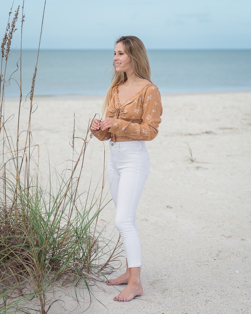 A young woman with long blonde hair, wearing a light brown floral top and white pants, stands barefoot on a sandy beach near tall grass, smiling and looking toward the ocean—an ideal moment for a Naples beach portrait photographer.