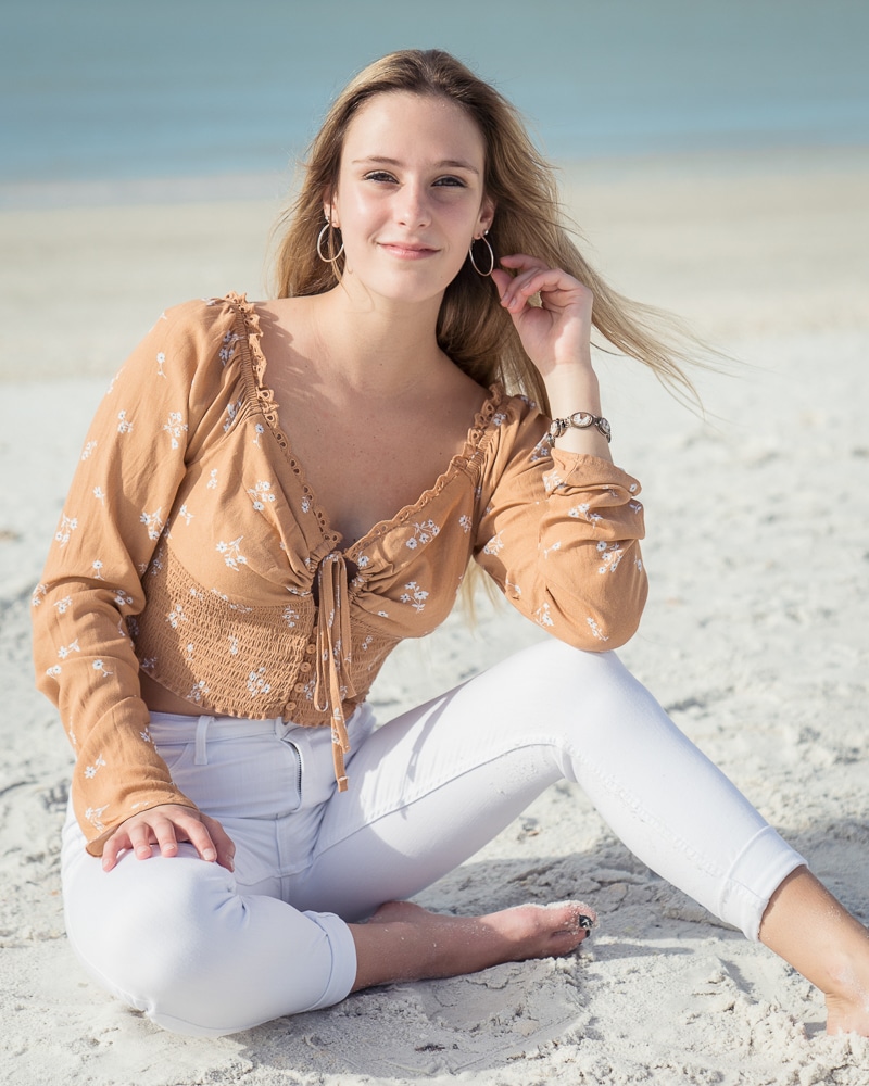 A young woman with long blonde hair sits on a sandy beach, wearing a light brown floral blouse and white pants. She smiles softly at the camera, with the ocean and sky in the background—captured by a Naples beach portrait photographer.